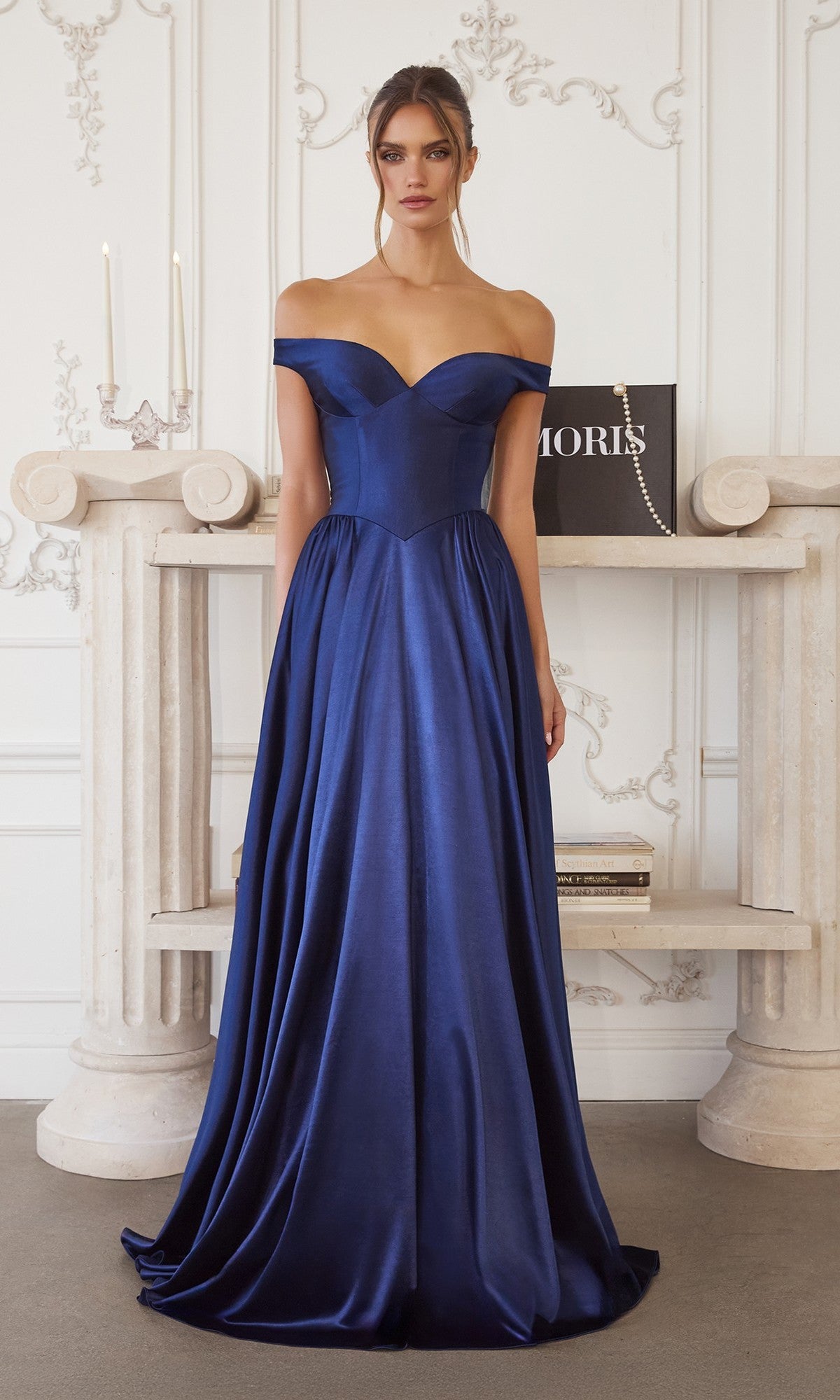 Indoors, a woman wears the Amoris T821 long prom dress—a navy off-the-shoulder satin gown with a fitted bodice and full A-line skirt. Ornate white wall paneling and a marble mantel appear in the background.